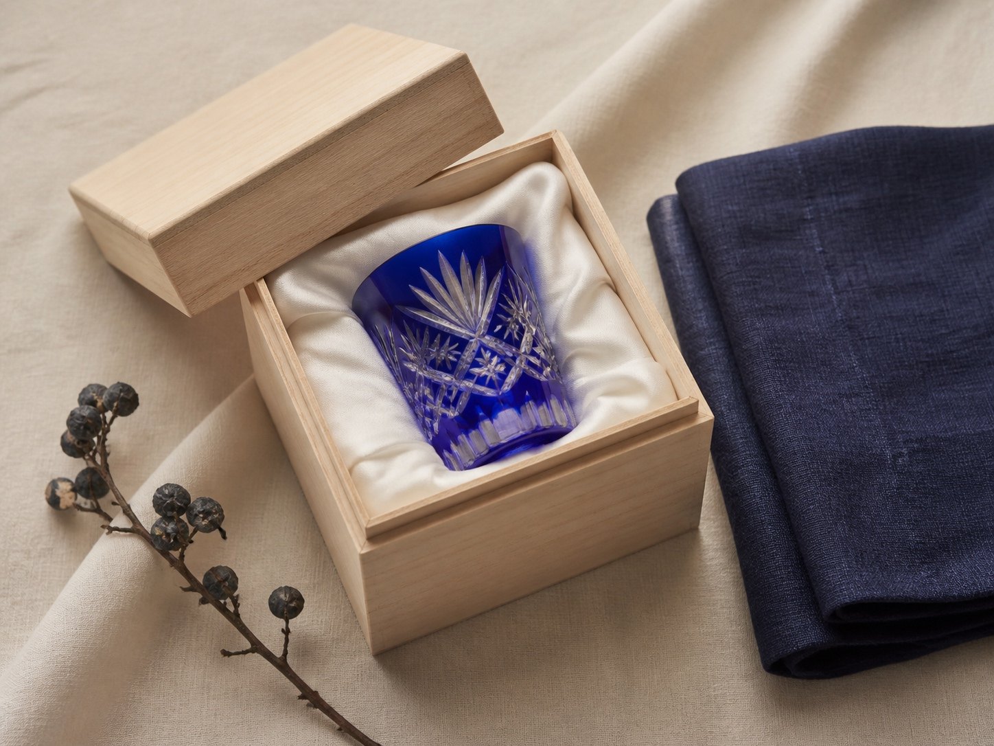 A blue Edo Kiriko sake cup resting in an opened paulownia wood gift box beside a folded indigo cloth