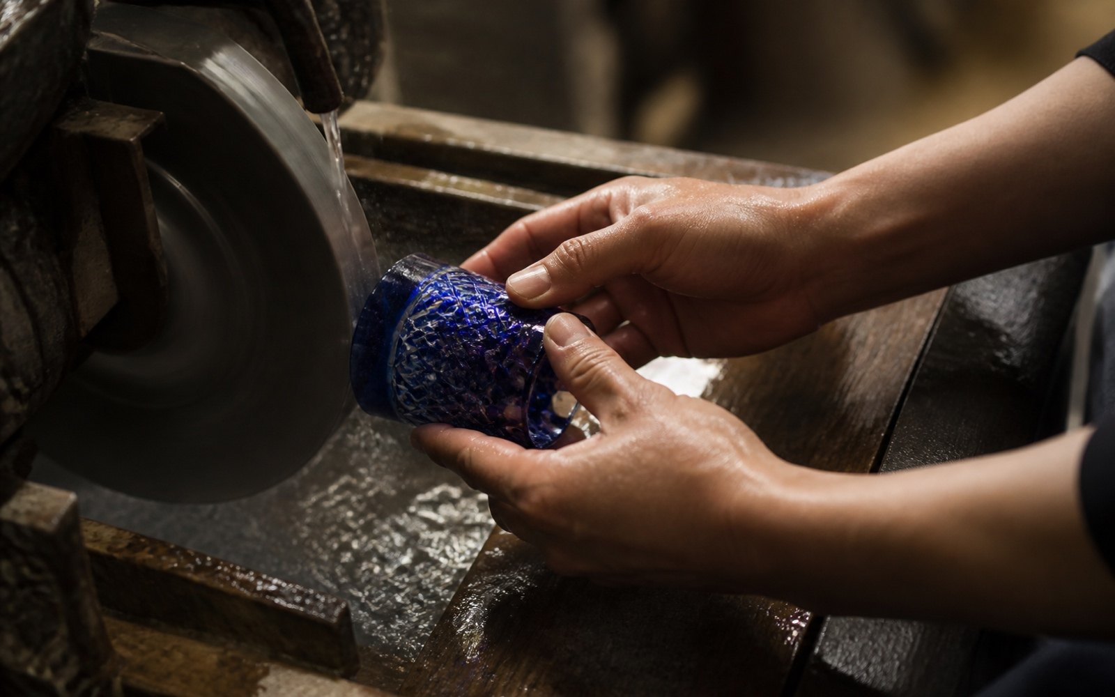 A craftsman's hands holding a cobalt blue glass against a spinning diamond wheel, cutting a facet into the colored surface